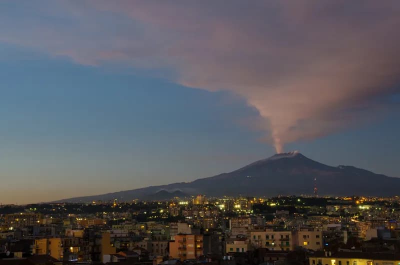 An ash plume rises from Mount Etna's summit above the lights of Catania at dusk.
