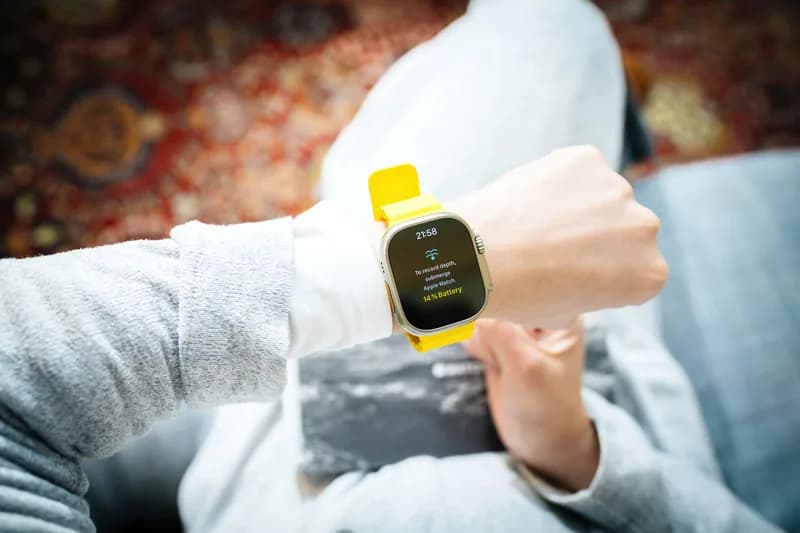 A yellow smartwatch worn on a wrist, with a hand reaching towards the screen.